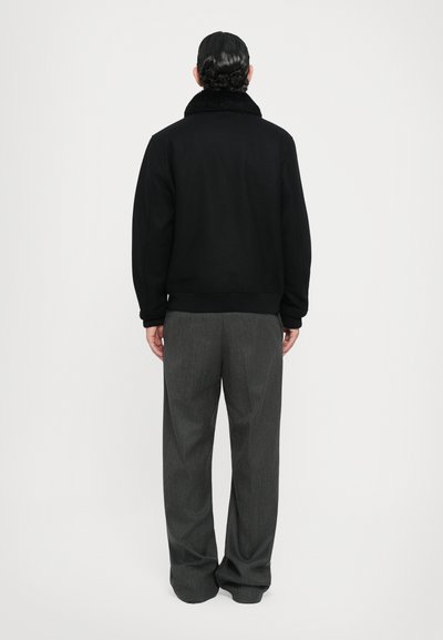 Schwarze Jacke mit Fellkragen, getragen mit grauen, weit geschnittenen Hosen. Glatte Textur und lockere Passform aus der Rückansicht sichtbar.