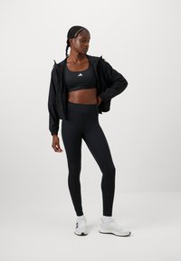 Fekete sportos ruházati szett tartalmaz egy crop topot és magas derekú leggingset, amelyet egy fekete könnyű dzseki és fehér sportcipői egészítenek ki.