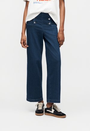 Άτομο που φοράει σκούρο μπλε τζιν παντελόνι με φαρδιά πόδια, με διπλό κουμπί μπροστά και μαύρα αθλητικά παπούτσια με λευκά κορδόνια και σόλα.