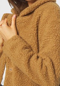 Brązowa kurtka polarowa z teksturowaną, puszystą powierzchnią. Wyposażona w kaptur i zapięcie na guziki z boku. Miękki, ciepły materiał.