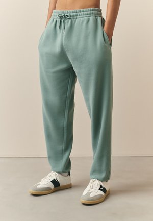 Osoba oblečená v tyrkysových teplákoch so šnúrkou a bielych teniskách s čiernymi pruhmi, stojaca s rukami v vreckách na svetlej podlahe.