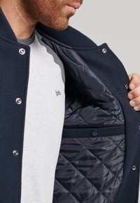 Marinblå quiltad jacka med silverfärgade tryckknappar, ribbad krage och slät innervadding. Grå T-shirt under.