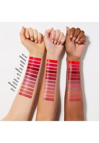 Trois bras avec différentes teintes de peau montrant douze échantillons de nuances de rouge à lèvres rouges et roses disposés verticalement sur chaque bras.