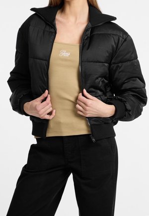 Femme portant une doudoune noire par-dessus un débardeur côtelé beige avec le logo "Guess Jeans", associée à un pantalon noir sur fond blanc.