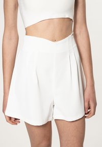 Pantaloni bianchi con vita alta, caratterizzati da pieghe e una superficie liscia. L'orlo è arrotondato, con una finitura pulita. Il tessuto è leggero e compatto.