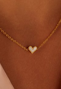 Gouden ketting met een klein hartje hanger met een witte emaille inlay en een gouden rand, rustend tegen een bruine huid.