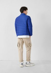 Man in een blauwe, gestikte jas, beige cargobroek, witte sokken en grijze sneakers, staand met zijn gezicht naar een witte muur en met zijn handen in zijn zakken.
