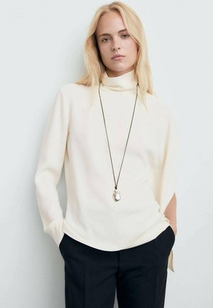 Maglione a collo alto color crema con maniche lunghe, realizzato in un tessuto morbido e con una vestibilità rilassata, abbinato a pantaloni scuri e un collier con pendente.