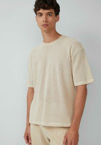 Beige gebreide t-shirt met korte mouwen, een losse, ademende textuur en een subtiel open geometrisch patroon. Ronde halsontwerp.