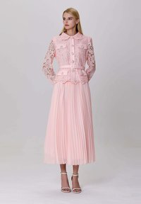 Femme portant une veste en dentelle rose pâle avec des boutons en perle et une taille ceinturée, associée à une jupe midi plissée assortie et des sandales à talons blancs.