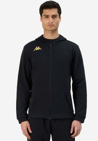 Felpa nera con zip realizzata in tessuto morbido, dotata di tasca a marsupio, polsini a coste e un logo dorato sul lato sinistro del torace.