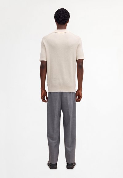 Uomo con capelli corti e ricci di spalle, indossa una maglietta beige a maniche corte in maglia, pantaloni grigi e scarpe nere su sfondo bianco.