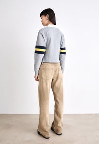 Felpa grigia corta con maniche a righe navy e gialle, abbinata a pantaloni beige a gamba larga e scarpe nere. Texture semplice e morbida.