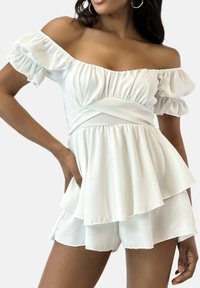 Witte off-shoulder peplum top met gerafelde mouwen en een gelaagde zoom, gecombineerd met witte shorts. Zachte, lichte stof met subtiele textuur.