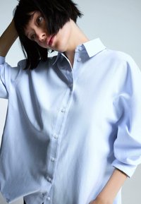 Jasnoniebieska, oversize koszula wykonana z gładkiego materiału, z kołnierzykiem w kształcie spiczastym, zapięciem na guziki z przodu i luźnymi rękawami o długości trzech ćwierci.