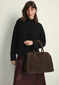 Brązowa zamszowa torebka z podwójnymi uchwytami, lekko usztywnionym kształtem i zamkiem błyskawicznym, trzymana przez osobę ubraną w czarny sweter i bordową spódnicę.