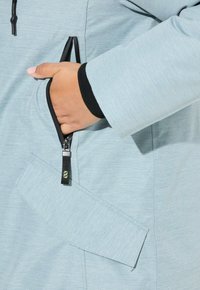Giacca azzurra chiara con tessuto testurizzato, zip nera e rifiniture, dotata di tasca laterale e un design di tasca angolare unico.