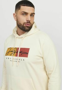 Krém színű kapucnis pulcsi puha anyagból, nagy piros és sárga grafikával, rajta a "Jack & Jones" és az "Originals Worldwide" felirattal.