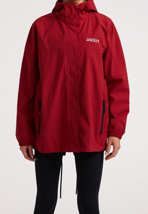Osoba ubrana w czerwony kurtkę z kapturem, z kieszeniami na zamek i logo "JACK1T" na piersi, w czarnych spodniach, stojąca na białym tle.
