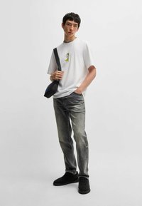 Hvid T-shirt med gul grafik, parret med grå jeans. Modellen bærer en sort crossbody taske og sorte sneakers. Simpel, afslappet påklædning.