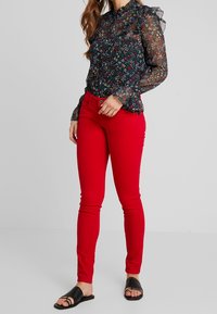 Vaqueros ajustados rojos combinados con una blusa negra de tul floral con mangas rizadas. La modelo lleva sandalias negras con tiras.