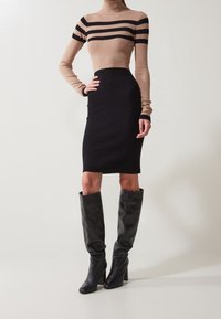 Donna in un vestito a collo alto a strisce beige e nere, abbinato a stivali alti in pelle nera, in piedi su uno sfondo neutro.