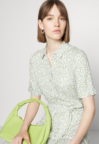 Vestido camiseiro de padrão floral em verde claro, com mangas curtas e cinto na cintura, combinado com uma bolsa redonda de cor verde brilhante.