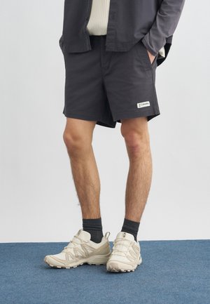 Άνδρας που φοράει μαύρο σορτς, σκούρες κάλτσες, μπεζ αθλητικά παπούτσια και σκούρο πουκάμισο με τα χέρια στις τσέπες, στέκεται σε μπλε χαλί.