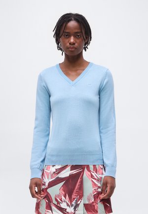 Žena s krátkymi copy, oblečená v svetlomodrom svetri s výstrihom do V a sukni s červenými a bielymi tropickými listovými vzormi.