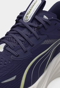 Ναυτικό μπλε παπούτσι για τρέξιμο με κορδόνια με σχέδιο, ανώτερο μέρος από δίχτυ, λευκό και κίτρινο λογότυπο Puma και παχύ λευκό πέλμα.