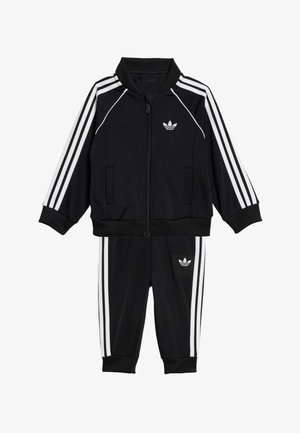 Mustan värinen verryttelypuku, johon kuuluu vetoketjullinen takki korkealla kauluksella ja valkoisilla raidoilla sekä samasta sileästä kankaasta valmistetut housut.