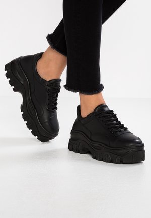 Osoba nosząca czarne masywne sneakersy na platformie i czarne spodnie z postrzępionym dołem, stojąca na białej powierzchni na jednolitym tle.