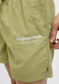 Svetlo olivové kraťasy vyrobené z ľahkého materiálu, s bočnými vreckami a potlačeným logom "Originals Studio" v bielej farbe.