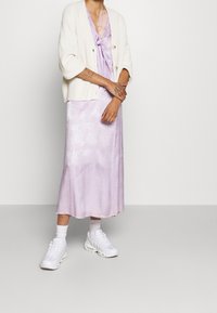 Hellviolettes Satinkleid mit einer Schleifendetail, darunter ein weißer, gerippter Cardigan. Kombiniert mit weißen Turnschuhen und Knöchelsocken.