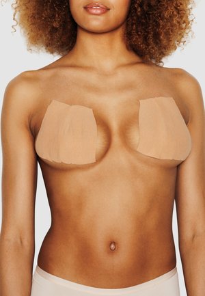 Donna con capelli ricci che indossa copriseno adesivi beige sul petto nudo su sfondo uniforme.