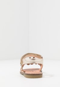 Dětské sandály s metalickým páskem a suchým zipem, plochou podešví a polstrovanou stélkou, zobrazené zezadu na bílém povrchu.