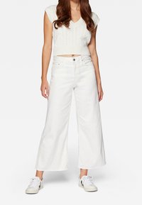 Nainen, joka pukeutuu valkoiseen, hihattomaan neulotun topin ja leveälahkeisiin valkoisiin farkkuihin sekä valkoisiin lenkkareihin mustilla yksityiskohdilla, seisoo yksinkertaisella taustalla.