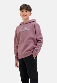 Garçon souriant portant un sweat à capuche rose avec le logo "Jack & Jones", associé à un pantalon cargo noir, se tenant avec confiance devant un fond uni.
