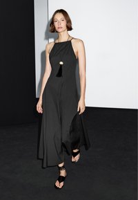 Černé bezpředělové šaty s splývavým designem a vysokým výstřihem. Obsahují dlouhý černý třásňový přívěsek. Nosené s černými sandály na tenkých řemíncích.
