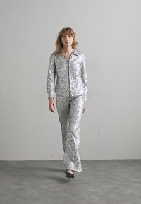 Sølvpaljetts pyjamas sett med glidelås foran jakke og lange bukser, med en jevn tekstur og løs passform. Svarte sko fullfører looken.