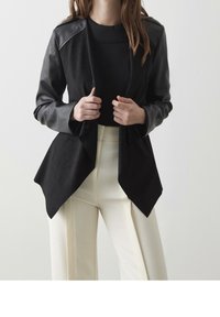 Veste en cuir synthétique noire avec une finition texturée, ourlet asymétrique et col rabattu, associée à un pantalon large crème.
