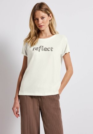 Short-sleeve cream t-shirt with rounded neckline, featuring black textual print "reflect" in a playful font, paired with textured brown pants.
