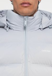 Light gray puffer jacket with a high collar, zip closure, and Calvin Klein logo, featuring a quilted design and smooth texture.