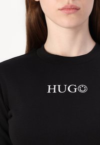 Crna rebrasta majica s bijelim tiskanim logotipom "HUGO" i grafikom nasmijanog lica, okrugli dekolte i pripijeni kroj.