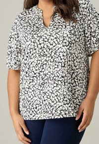 Blusa de estampado floral en blanco y negro con mangas cortas, escote en pico y un corte relajado, combinada con pantalones de color azul oscuro.