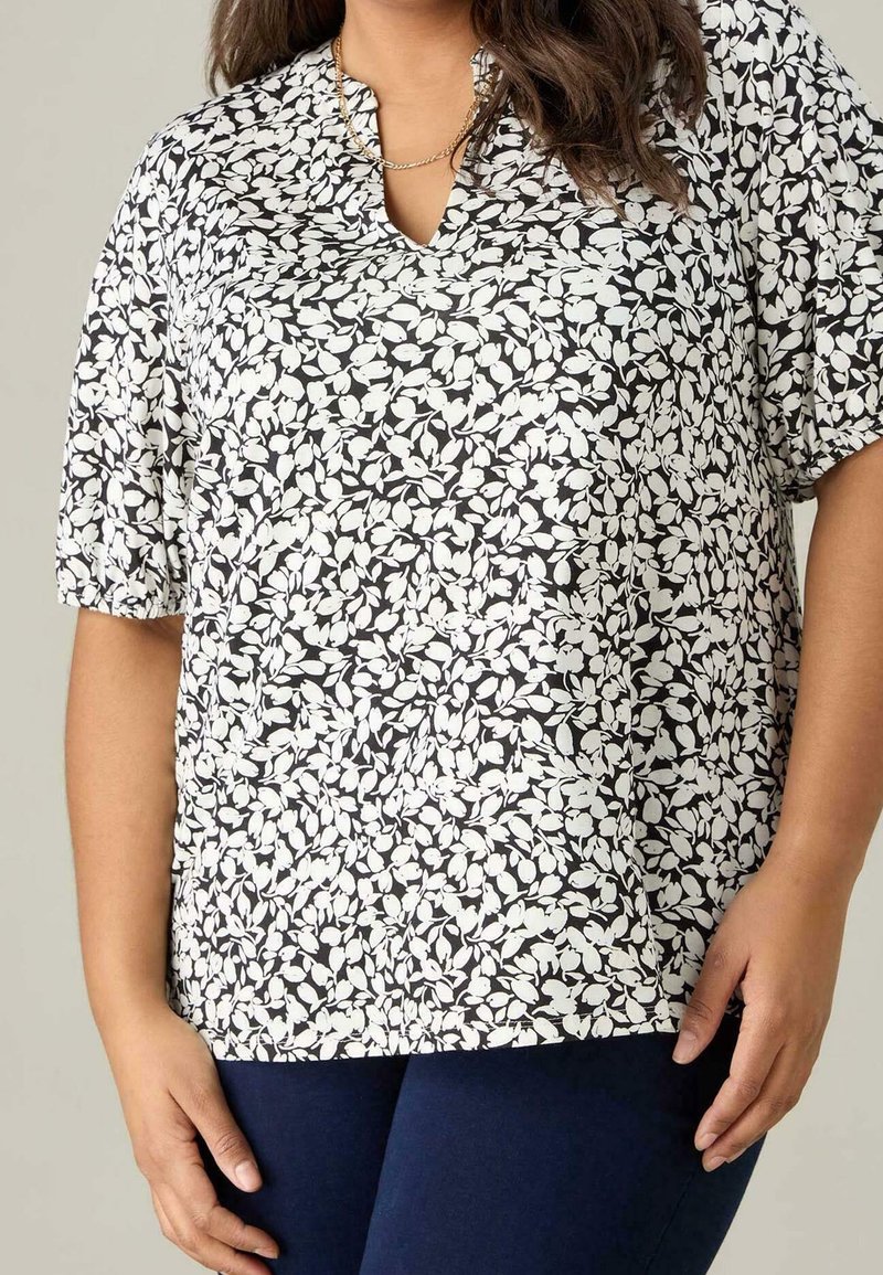 Blusa de estampado floral en blanco y negro con mangas cortas, escote en pico y un corte relajado, combinada con pantalones de color azul oscuro.