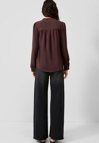 Vrouw die met de rug naar de camera staat, gekleed in een bordeauxrode blouse met lange mouwen en zwarte, wijd uitlopende jeans, tegen een lichte, effen achtergrond.