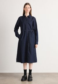 Denimskjortklänning med knappstängning fram, långa ärmar, midjeband, sidofickor och knälång fåll i mörkblått. Svarta platåskor bärs.