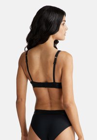 Žena s dlouhými tmavými vlasy, oblečená v černé podprsence a černém spodním prádle, zobrazená ze zadu na jednolitém bílém pozadí.