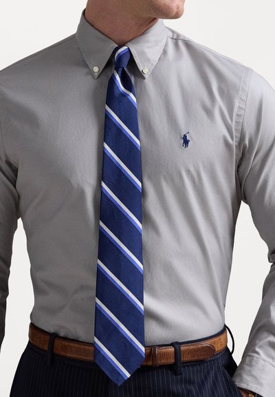 Camicia grigia con colletto button-down e cravatta blu navy con strisce diagonali azzurre, abbinata a una cintura marrone e pantaloni blu navy a righe.